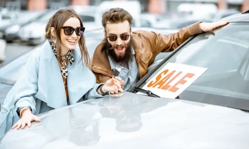 Deux personnes souriantes devant une voiture avec panneau SALE illustrant l’achat aux enchères d’un véhicule d’occasion