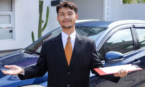 Homme en costume noir avec cravate orange tenant un dossier rouge devant une voiture bleue illustrant une présentation liée au rachat de soulte