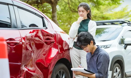 Un homme accroupi commence à remplir un constat amiable sur papier devant une voiture rouge accidentée tandis qu'une femme observe les dégâts avec inquiétude
