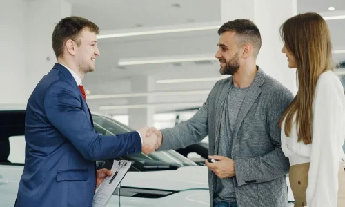 Client en costume serrant la main d’un conseiller près d’une voiture, image illustrant le rappel constructeur et la procédure à suivre pour vérifier son véhicule.