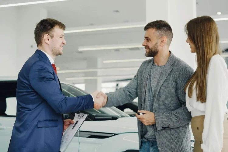 Client en costume serrant la main d’un conseiller près d’une voiture, image illustrant le rappel constructeur et la procédure à suivre pour vérifier son véhicule.