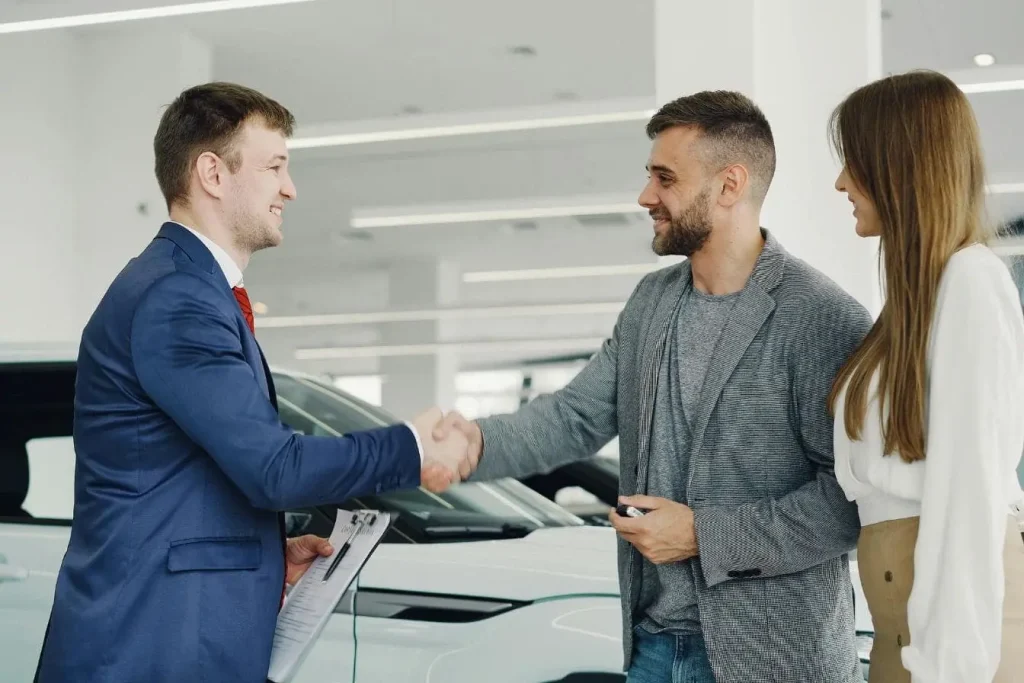 Client en costume serrant la main d’un conseiller devant voiture blanche, image illustrant la confiance et la protection offertes par la garantie constructeur automobile.