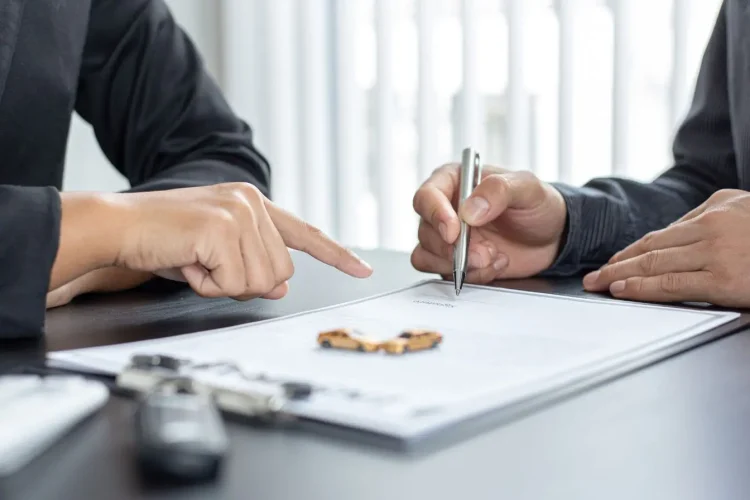Deux personnes assises signant un document avec clés et voitures miniatures sur table, image illustrant la souscription d’une assurance auto tous risques.