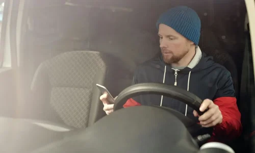 Personne portant bonnet bleu et veste bicolore, tenant un téléphone en conduisant, symbole des risques liés au téléphone au volant.