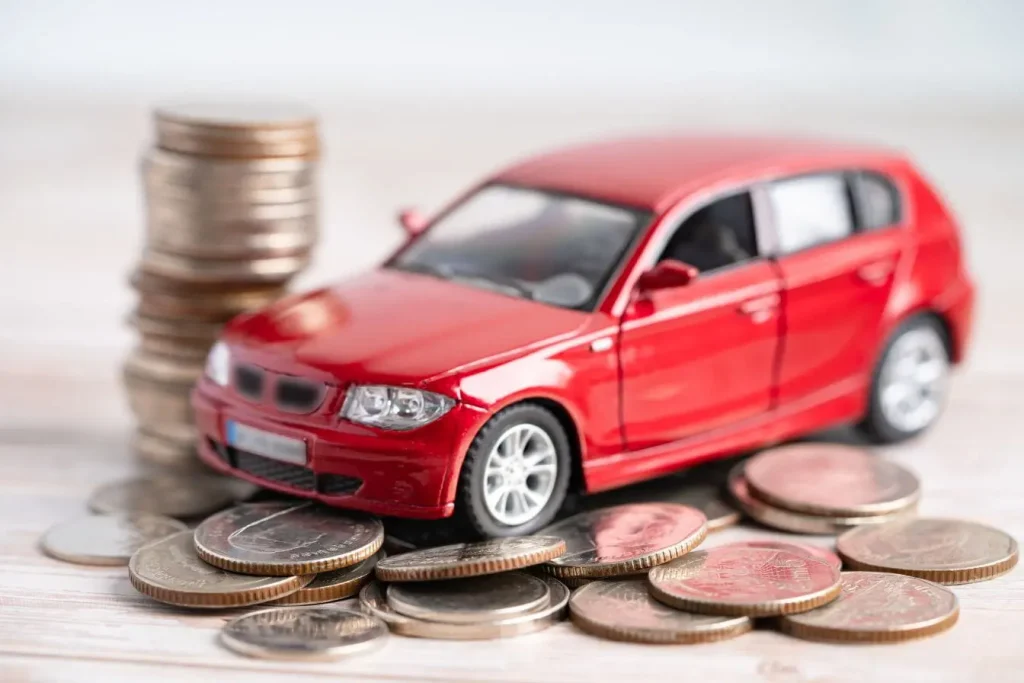 Voiture miniature rouge entourée de pièces empilées sur bois, illustrant la responsabilité civile automobile et les coûts liés à la protection.