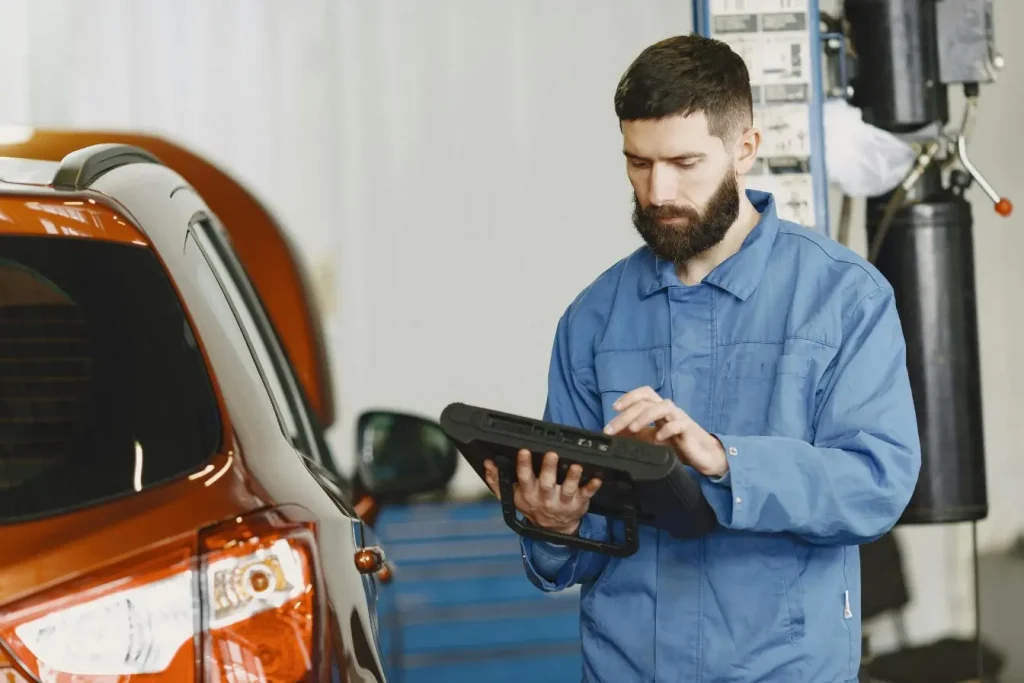 Mécanicien en bleu utilisant tablette électronique près d’une voiture orange, image illustrant le diagnostic OBD et l’analyse des systèmes embarqués.
