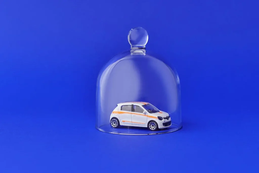 Voiture miniature blanche protégée sous une cloche en verre sur fond bleu, illustrant la responsabilité civile automobile et la notion de protection.