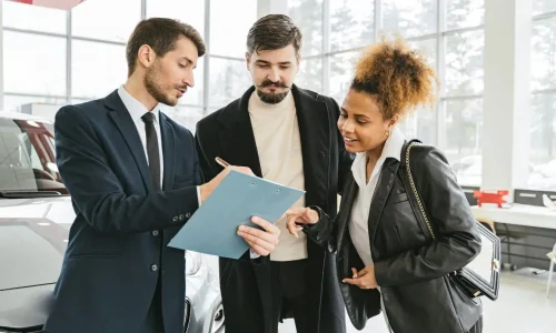 Trois personnes en showroom discutant devant une voiture argentée, image illustrant la signature d’un contrat d’assurance auto.