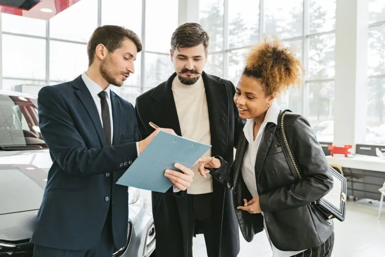 Trois personnes en showroom discutant devant une voiture argentée, image illustrant la signature d’un contrat d’assurance auto.