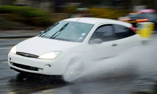 Voiture blanche roule vite sur route mouillée, éclaboussures massives, image claire du risque d’aquaplaning en conditions pluvieuses.