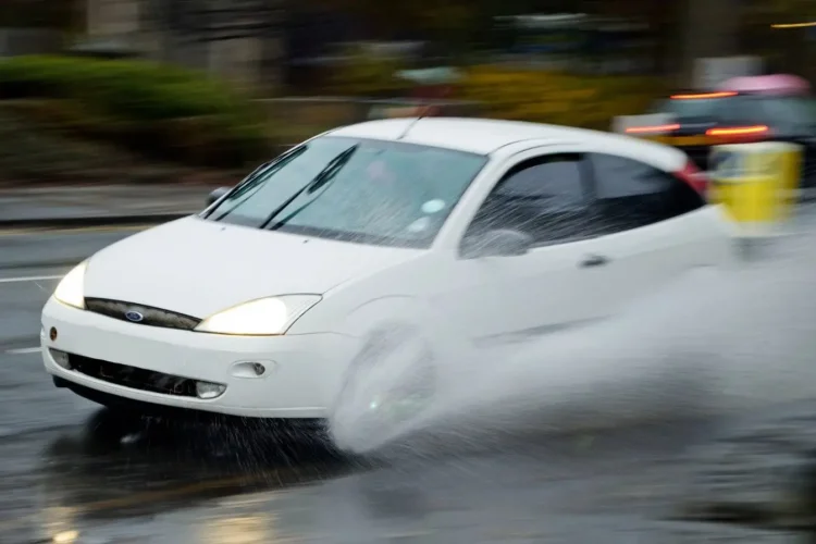 Voiture blanche roule vite sur route mouillée, éclaboussures massives, image claire du risque d’aquaplaning en conditions pluvieuses.