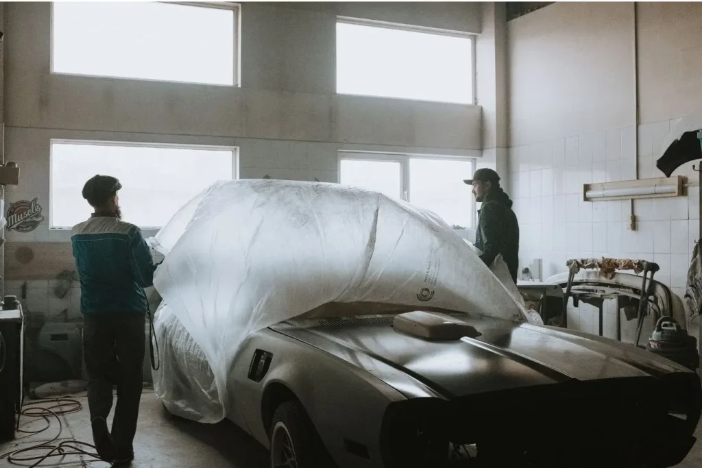 Deux personnes retirant une bâche transparente sur une voiture vintage, image qui illustre la restauration minutieuse d’un modèle ancien.
