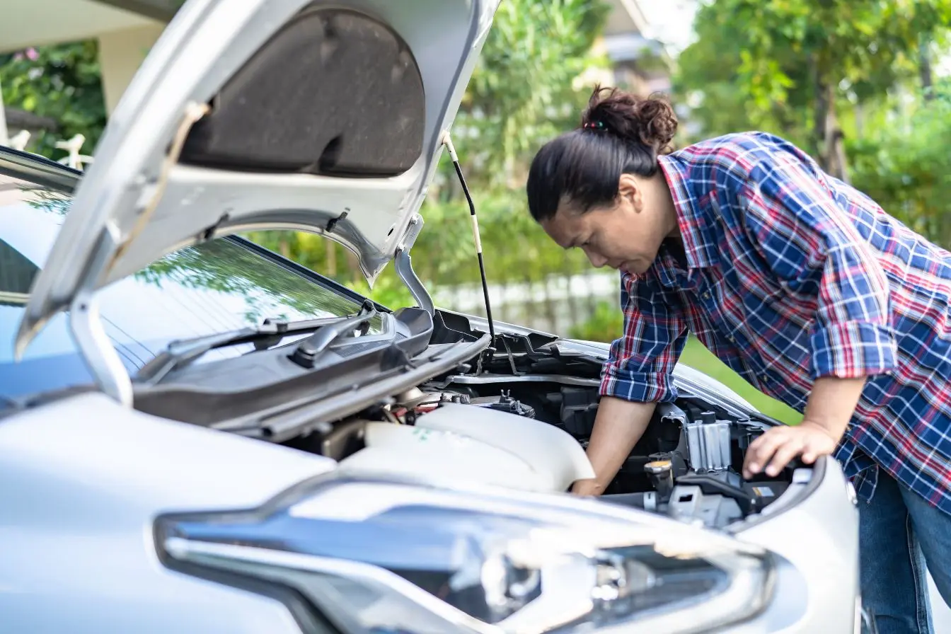 Une femme penchée sous le capot ouvert d'une voiture pour vérifier la mécanique et anticiper le prix moyen d'un changement de plaquettes de frein.