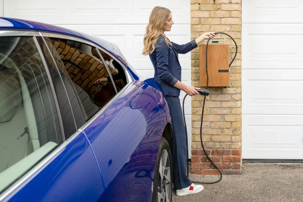 Personne tenant câble branché à voiture bleue devant garage, symbole du temps de charge sur prise domestique.